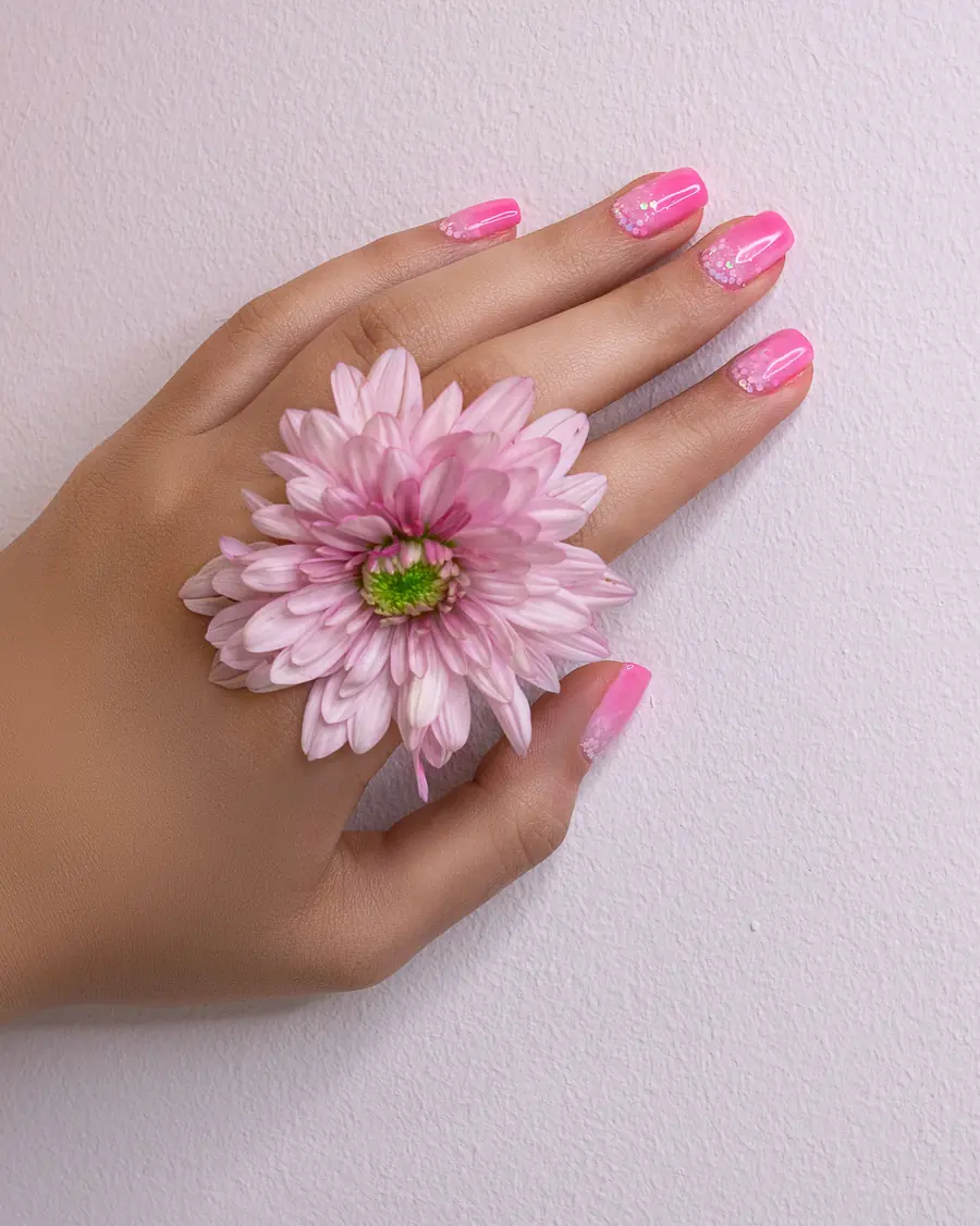 Main aux ongles roses tenant une fleur, sur un fond lilas clair